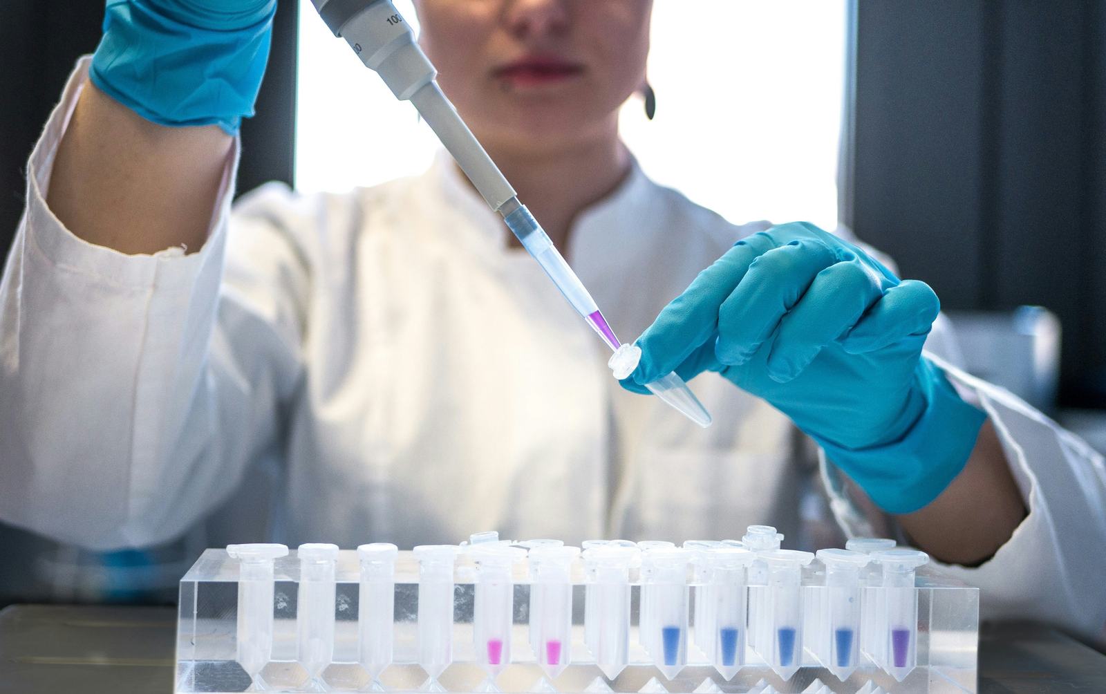 Technician in a lab coat and gloves pipetting samples into a rack of tubes